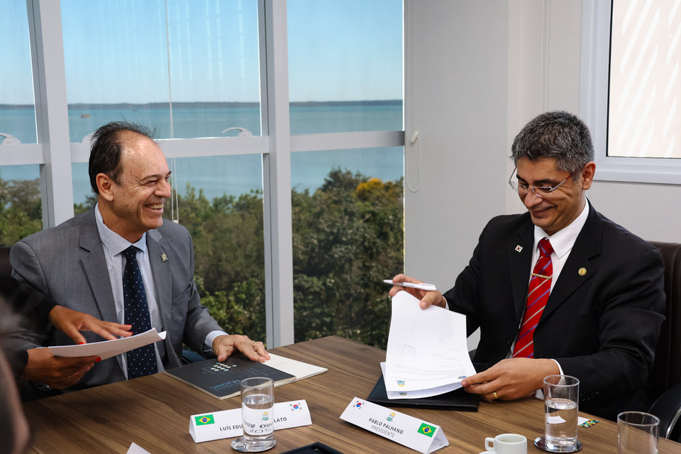Homem branco sorrindo ao lado de homem amarelo assinando documento